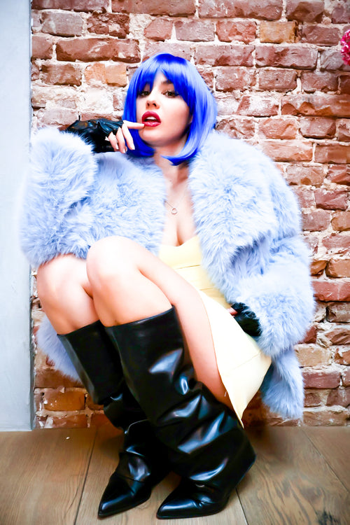 Person with blue hair and a fluffy blue coat sitting against a brick wall.