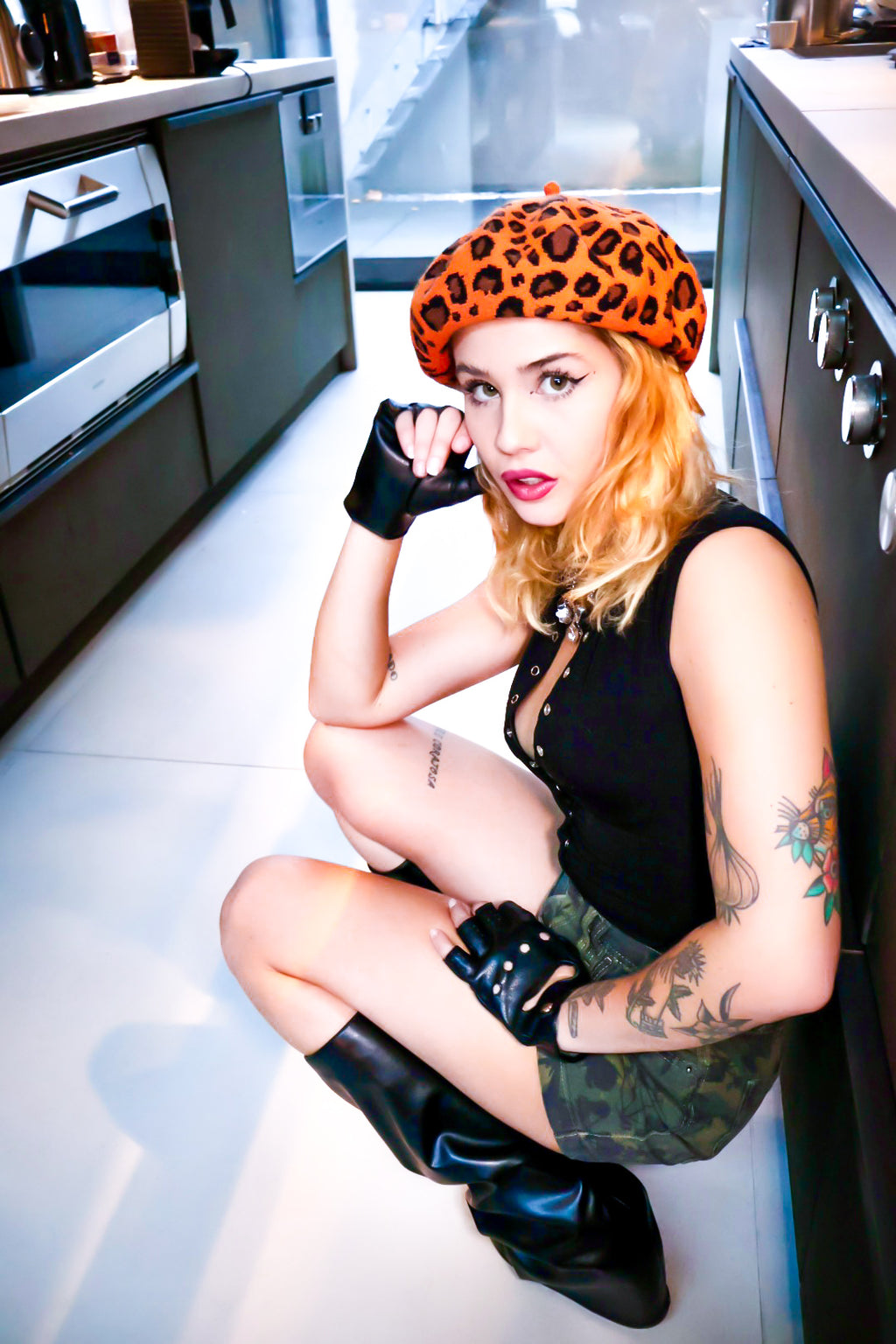Woman in leopard print hat and black gloves sitting on a kitchen floor.