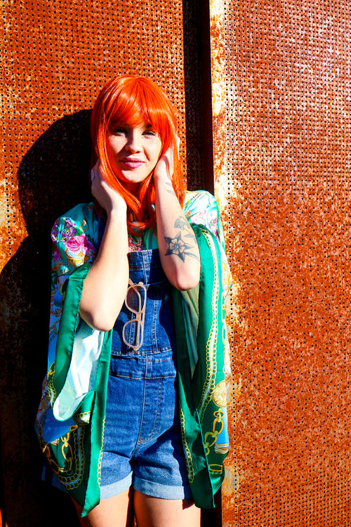 Person with vibrant red hair wearing a colorful kimono-style garment against an orange textured wall.