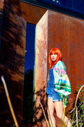 Person with bright red hair wearing a floral kimono standing in front of a rusted metal wall.