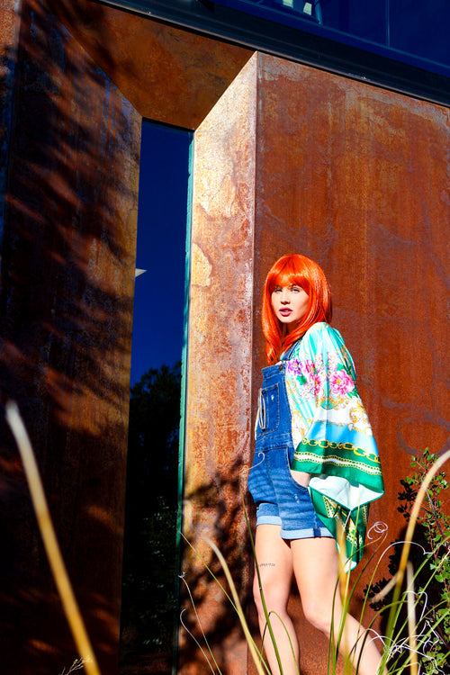 Person with bright red hair wearing a floral kimono standing in front of a rusted metal wall.
